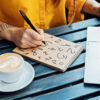 mulher sentada apoiada numa mesa de madeira com um bloco escrevendo números o notebook e uma xícara de café