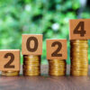 o número 2024 está escrito em blocos de madeira que estão em cima de pilhas de moedas com um fundo de folhas verdes desfocadas