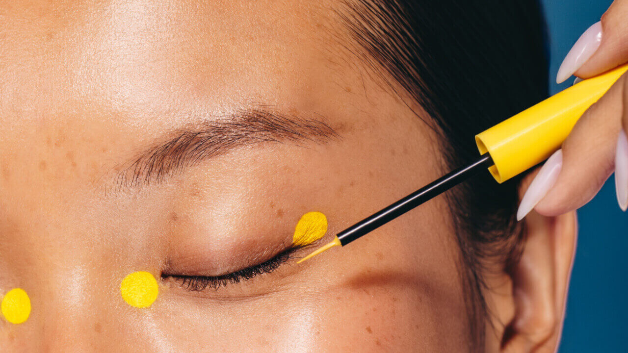 Mulher fazendo maquiagem com um produto cosmético, aplicando quatro pontos ao redor dos olhos usando um delineador amarelo, uma técnica de maquiagem.