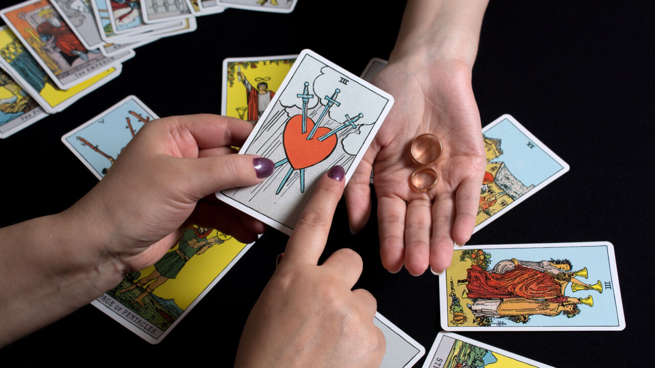 em cima de uma mesa com cartas de tarot uma mão branca segura uma carta de tarot com um coração e ao lado há uma mão segurando duas alianças