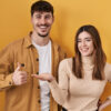 à esquerda está um homem branco e fazendo sinal de positivo com o dedo e à direita uma mulher com a palma da mão esticada eles estão em fundo amarelo e com roupas nesta mesma paleta de cores
