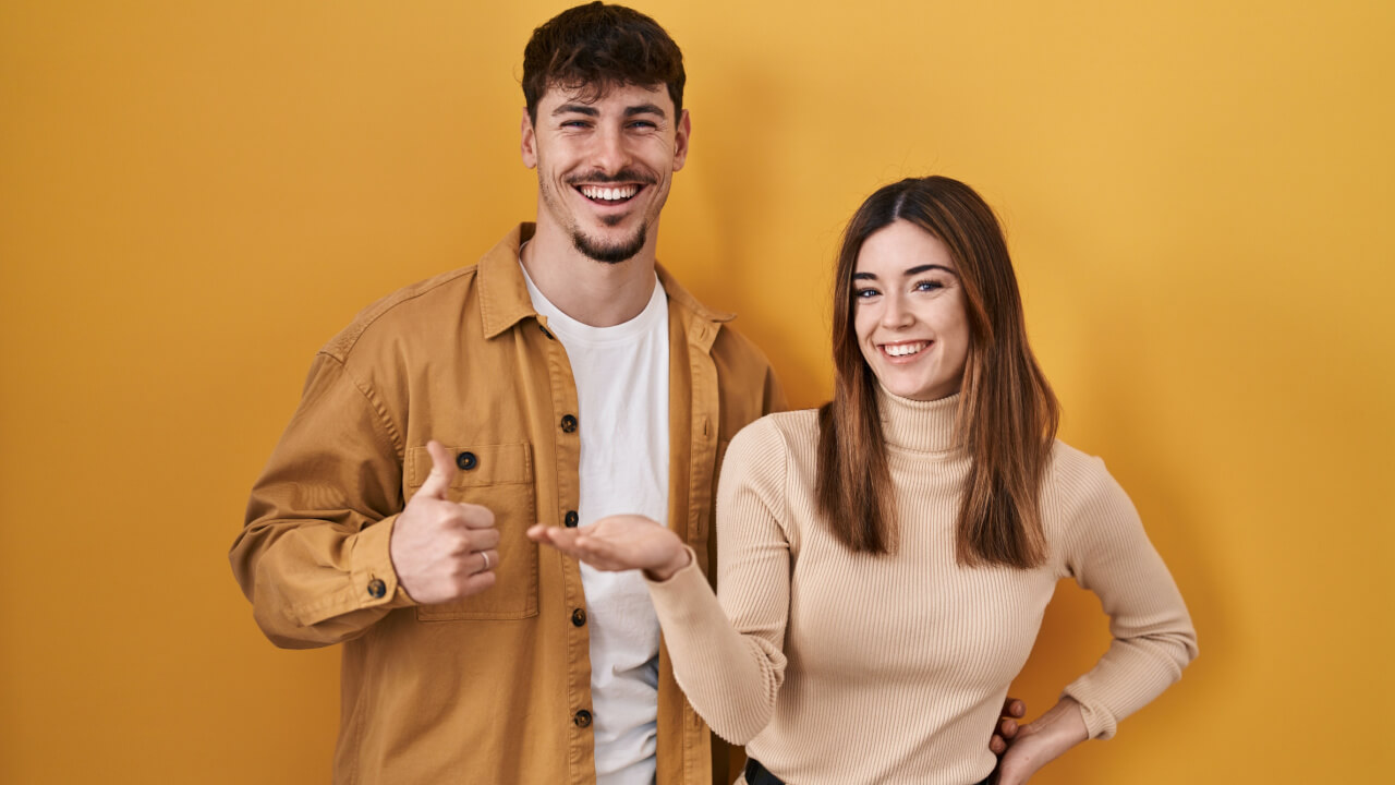 à esquerda está um homem branco e fazendo sinal de positivo com o dedo e à direita uma mulher com a palma da mão esticada eles estão em fundo amarelo e com roupas nesta mesma paleta de cores