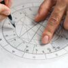 mão fazendo um desenho a caneta sobre um papel branco com uma mandala astrologica desenhada