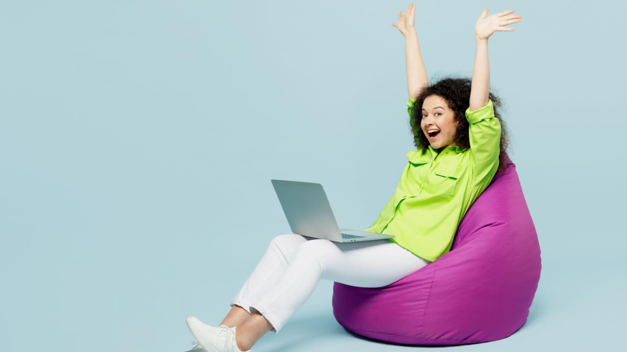 Foto de corpo inteiro de uma mulher usando uma camisa verde casual, sentada em um pufe, com um laptop no colo, com os braços levantados, isolada em um fundo azul pastel. Conceito de estilo de vida.