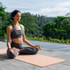 imagem de mulher sentada num tapete de yoga fazendo meditação ao ar livre