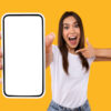 mulher branca com cabelo liso e segurando um celular e apontando para ele em fundo amarelo