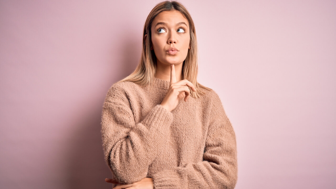 Mulher loira usando um suéter de lã sobre um fundo rosa, isolada, pensando, concentrada, com o dedo no queixo, olhando para cima.