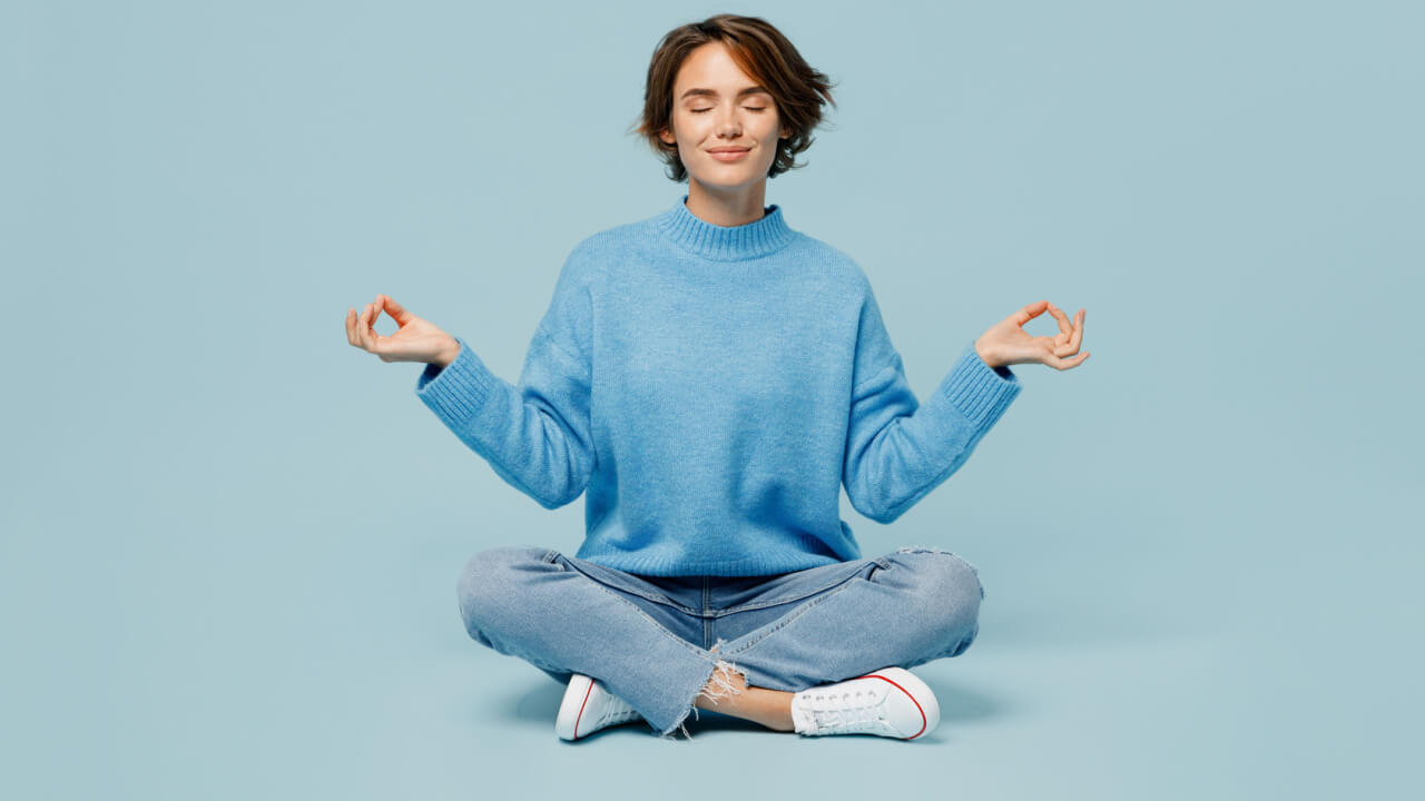 Foto de corpo inteiro de uma mulher usando um suéter, com as mãos em gesto de ioga, relaxando, meditando, tentando se acalmar, isolada em um fundo azul pastel liso.