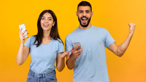 Uma mulher e um homem muito felizes, regozijando-se com o sucesso, segurando seus celulares, gritando e sorrindo com triunfo, de pé sobre um fundo amarelo.