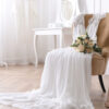 à direita está uma cadeira de cor marrom com um vestido de noiva em cima dela com um buquê e um sapato do lado dentro de uma sala grande com uma cortina branca ao fundo