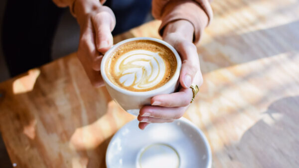 xícara branca com café onde a espuma forma uma flor e ao fundo há uma mesa de madeira