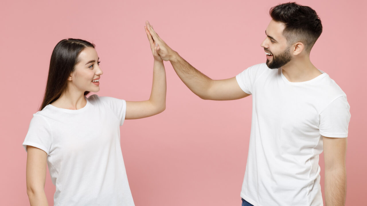 Casal (um homem e uma mulher) alegre, usando camisetas brancas básicas, dando um "high five", isolado em um fundo rosa pastel.