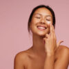 Mulher aplicando produto para a pele no rosto, sorrindo, contra um fundo rosa.