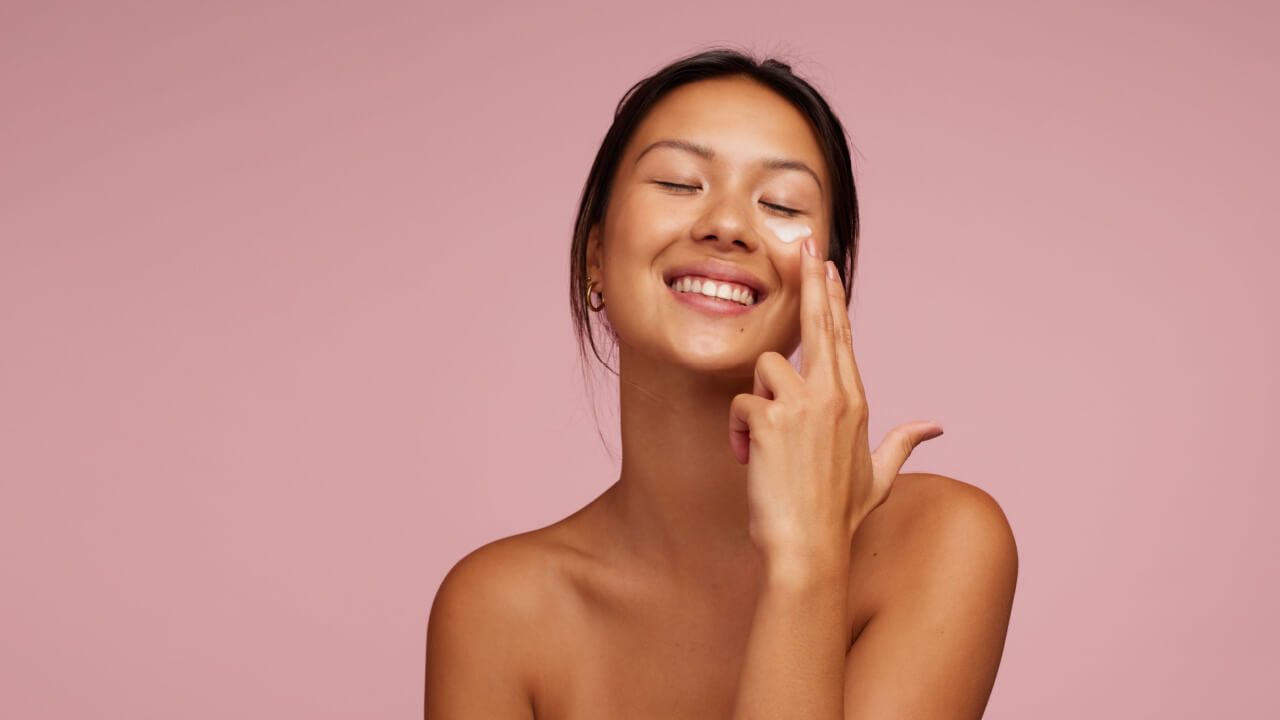 Mulher aplicando produto para a pele no rosto, sorrindo, contra um fundo rosa.
