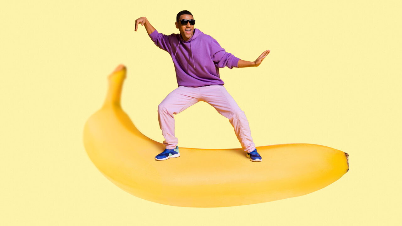Homem voando e dançando em cima de uma banana gigante, isolado em um fundo de cor amarelo pastel.