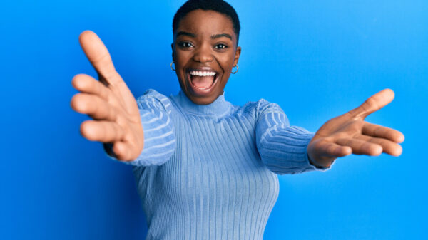 mulher negra de cabelos curtos sorrindo de braços abertos em fundo azul