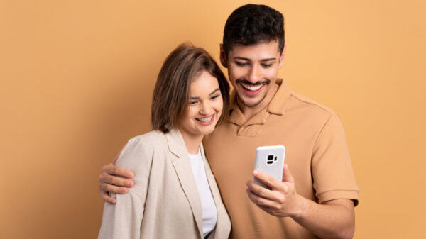 Casal alegre olhando para um celular, em um fundo de cor bege.