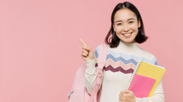 mulher de traços asiáticos carregando um caderno no braço esquerdo e uma mochila rosa clara pendurado no braço direito em fundo rosa