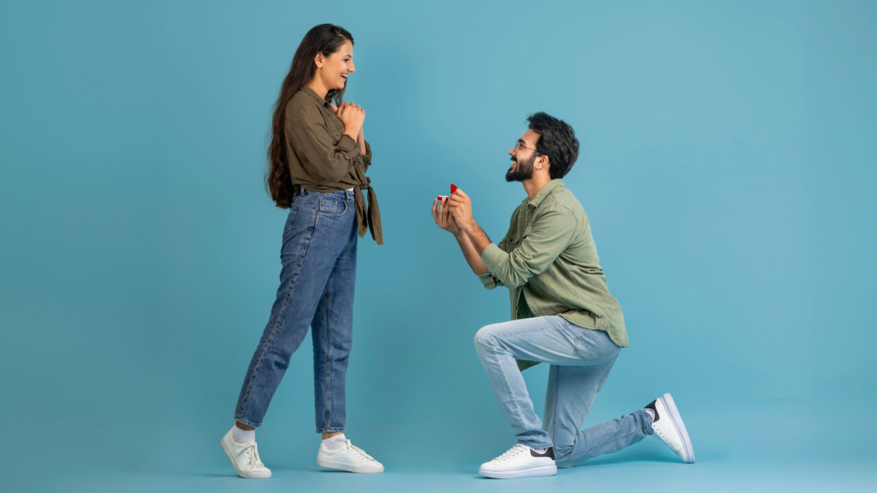 Homem feliz segurando uma caixa aberta com um anel de noivado para a namorada animada, pedindo que ela seja sua esposa, sobre um joelho, em uma parede azul de um estúdio.