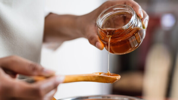 fotografia destacando um pote de vidro de mel em uma mão sendo despejado em uma colher de madeira que está em outra mão