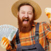 Homem barbudo usando um macacão e um chapéu de palha, segurando notas de dinheiro, fazendo gesto de vencedor, isolado em um estúdio de fundo de cor bege claro liso.