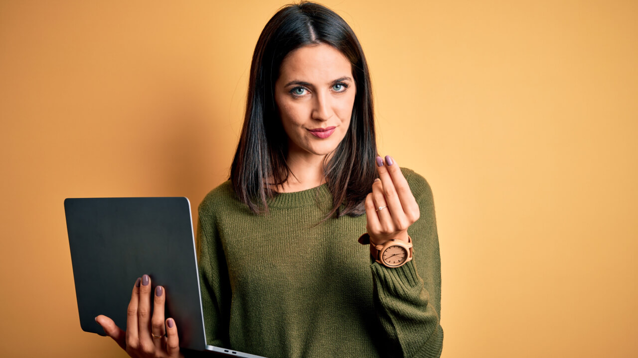 Mulher morena de olhos azuis usando um computador portátil sobre um fundo laranja, fazendo gesto de dinheiro com a mão.