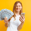 Retrato de uma mulher animada usando uma camiseta branca, mostrando notas de dinheiro e segurando um celular, isolada em um fundo amarelo.