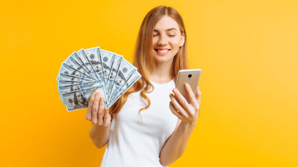 Retrato de uma mulher animada usando uma camiseta branca, mostrando notas de dinheiro e segurando um celular, isolada em um fundo amarelo.