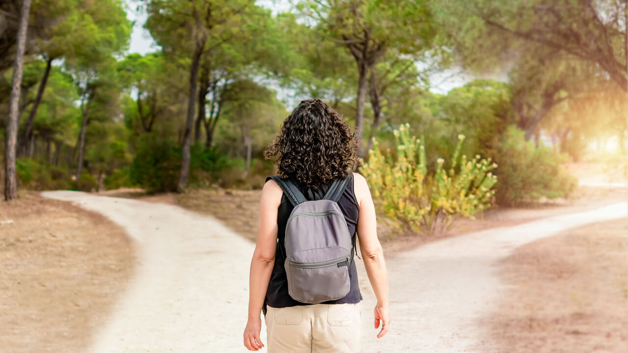 imagem de uma mulher de costas com uma mochila e ela está olhando para uma estrada de dois caminhos em uma floresta