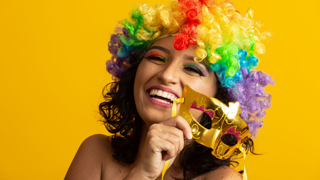 mulher sorrindo segurando uma mascara dourada de carnaval e uma peruca colorida em fundo amarelo