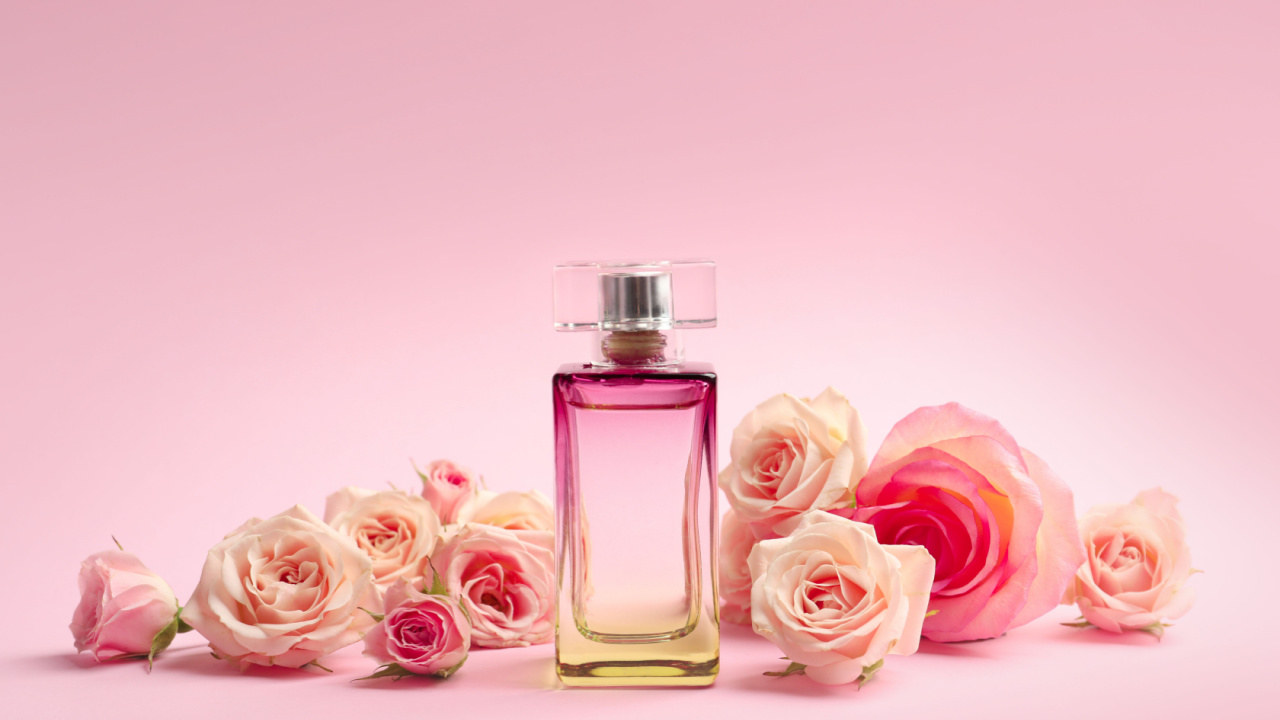 imagem com um vidro de perfume no centro e rosas aos lados em fundo rosa