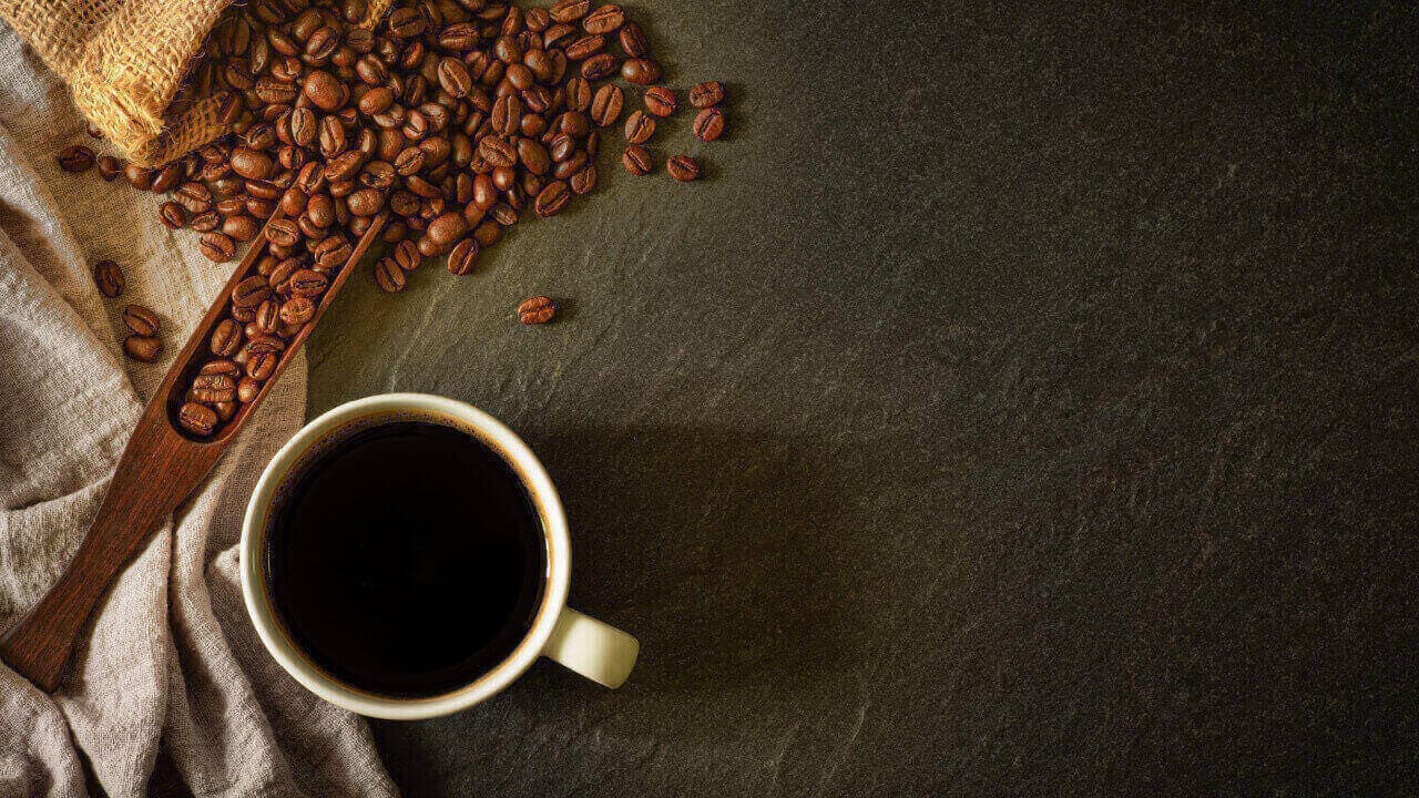 Café preto em uma xícara de cerâmica branca, com grãos de café em um saco sobre uma mesa escura. Vista superior, plana.