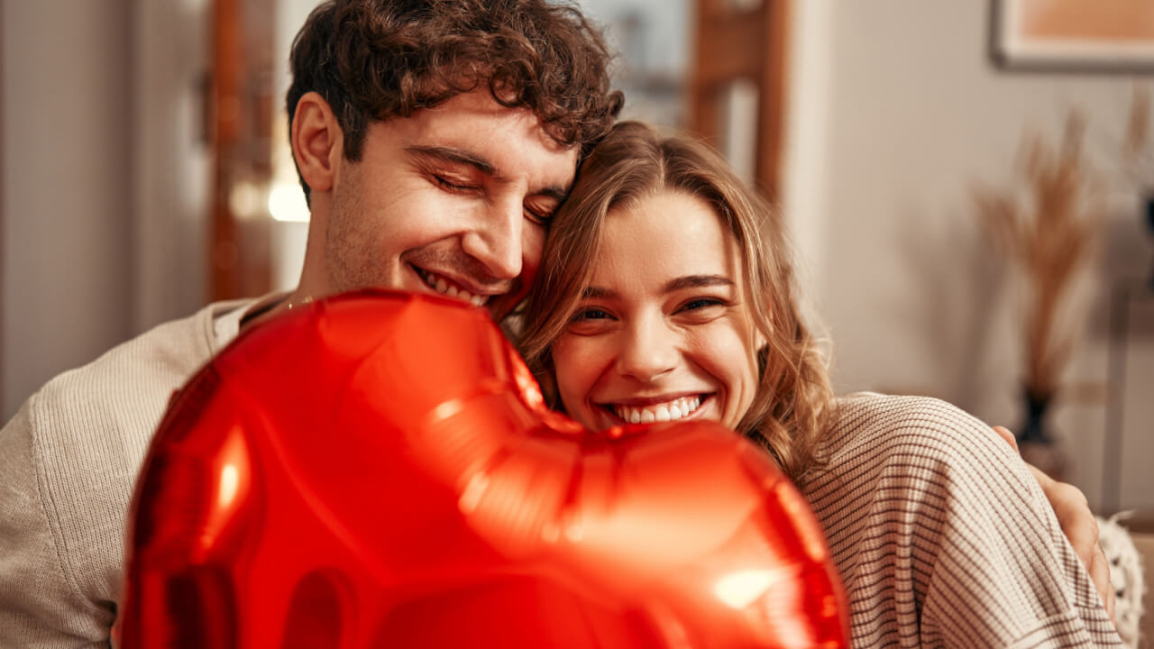 casal heterossexual juntos sorrindo e segurando um balão de coração na altura da boca