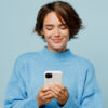 Mulher sorrindo, feliz, usando um suéter de malha, segurando um celular com as mãos, isolada em um retrato de estúdio de fundo azul claro liso.
