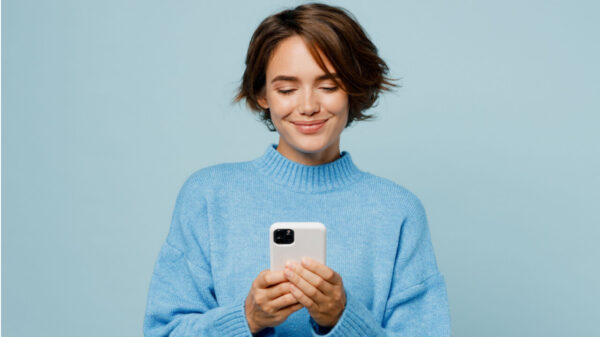 Mulher sorrindo, feliz, usando um suéter de malha, segurando um celular com as mãos, isolada em um retrato de estúdio de fundo azul claro liso.
