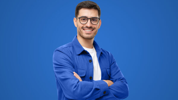 homem branco usando óculos e camisa azul. Ele está de braços cruzados em um fundo azul