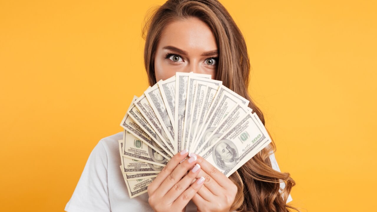 Retrato de uma jovem cobrindo o rosto com notas de dinheiro, isolada sobre um fundo amarelo.