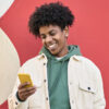 Homem feliz, sorrindo, em pé sobre uma parede vermelha, usando um celular e olhando para ele.