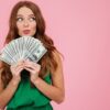 Retrato de uma mulher de cabelo longo segurando notas de dinheiro e olhando para o o lado, isolada sobre um fundo rosa.