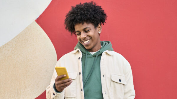Homem feliz, sorrindo, em pé sobre uma parede vermelha, usando um celular e olhando para ele.
