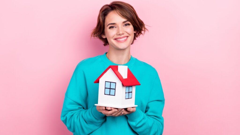 Retrato de uma garota alegre usando um pulôver, segurando uma casa em miniatura, isolada em um fundo de cor rosa.