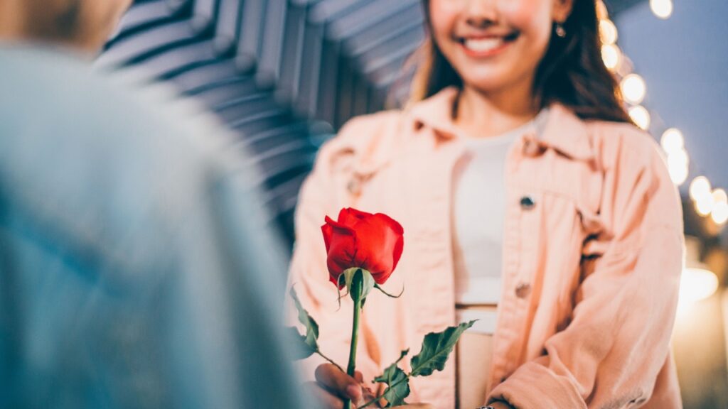 em primeiro plano está um botão de rosa vermelho. em segundo plano e desfocado está uma mulher sorrindo e usando uma jaqueta rosa claro recendo a flor de um homem ajoelhado
