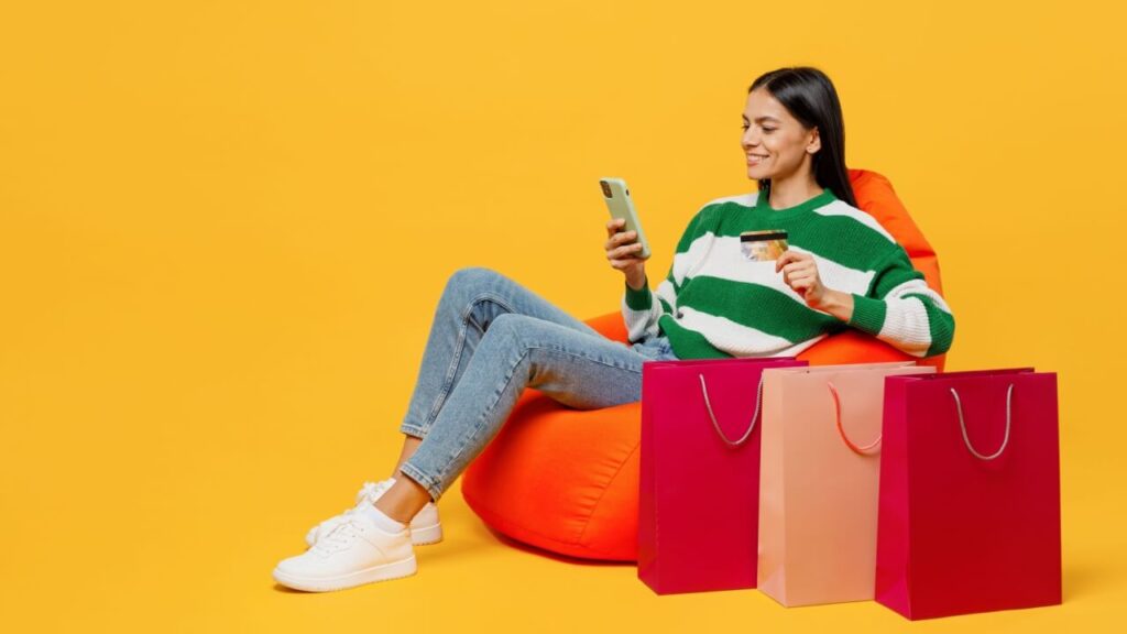 Jovem feliz usando um suéter de malha verde e branco, sentada em um pufe perto de sacolas de compra, usando um celular, isolada em um fundo amarelo liso. Conceito de compras.