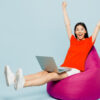 Jovem de corpo inteiro, usando uma camiseta vermelha casual, sentada em um pufe, usando um laptop, fazendo gesto de vencedor, isolada em um estúdio de fundo azul simples. Conceito de estilo de vida.