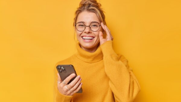 mulher vestindo roupa de frio amarela e segundo um celular na mão esquerda enquanto a mão direita está na altura dos olhos encostado no óculos em fundo amarelo