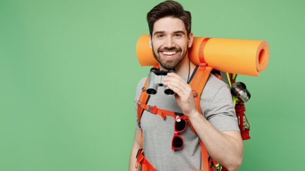 homem sorrindo com mochilas de viagem nas costas e segurando um binóculos em fundo verde