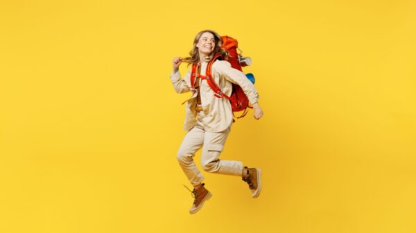 mulher pulando com uma mochila grande de viagem nas costas em fundo amarelo