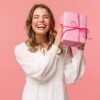 mulher sorrindo e segurando uma caixa de presente rosa em fundo também rosa