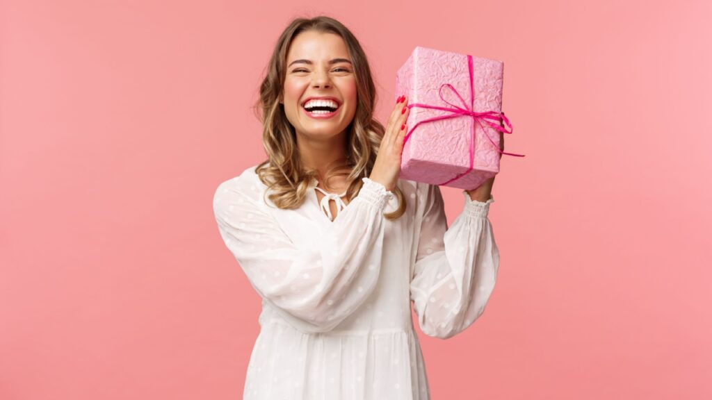 mulher sorrindo e segurando uma caixa de presente rosa em fundo também rosa