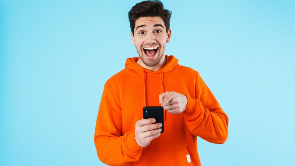 Imagem de um homem feliz e surpreso isolado sobre um fundo de parede azul, usando um celular.